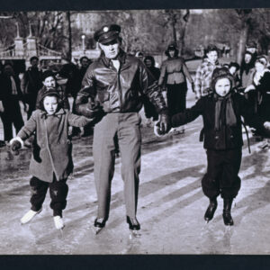 A black and white photograph of a White man in a winter military uniform of slacks, leather jacket, and brimmed hat holding the hand of a child on either side dressed in multiple layers of winter clothing. Behind the trio stands a group of adults and children with the grounds of Boston’s Public Garden behind them.