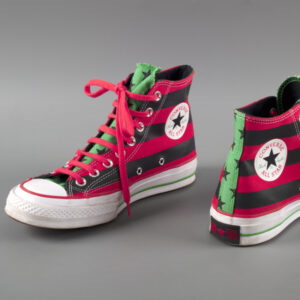 A pair of high-top canvas sneakers with one toe facing the viewer and the other with the heel facing the viewer. Main canvas top is made of thick, red and black, horizontal stripes, red shoelaces, round white Converse logo screen printed on sides, tongue and back heel strip are green fabric with five-pointed black stars. Soles are white rubber with green and red strips.