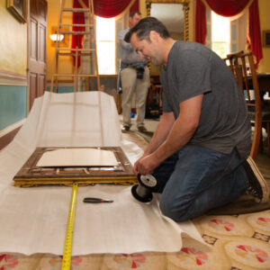 Photograph of a White man, with dark hair, and tanned skin wearing a plain, grey t-shirt, jeans, and black, high-top canvas sneakers kneeling in a historic home installing hanging wire to the back of a portrait frame.