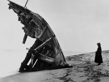 Black and white photo of a person wearing a long coat and hat standing in profile facing a worn down piece of a shipwreck on a beach.