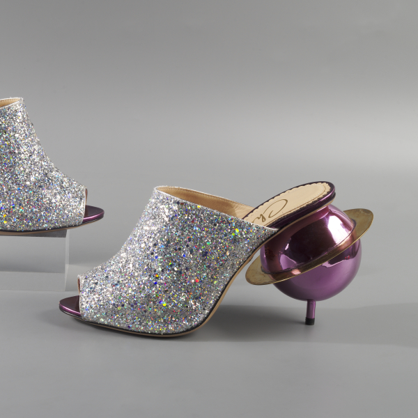A pair of sparkly silver mules with each heel in the shape of a pink Saturn with a large gold ring encircling it.