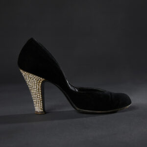 A side view of a high-heeled black velvet pump with clear rhinestones covering the back of the heel.