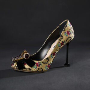 A side view of a high-heeled pump, with a thin black stem heel and patterned yellow and black fabric with green, purple, and red flowers. The toe features a black bow with a rhinestone clip at center.