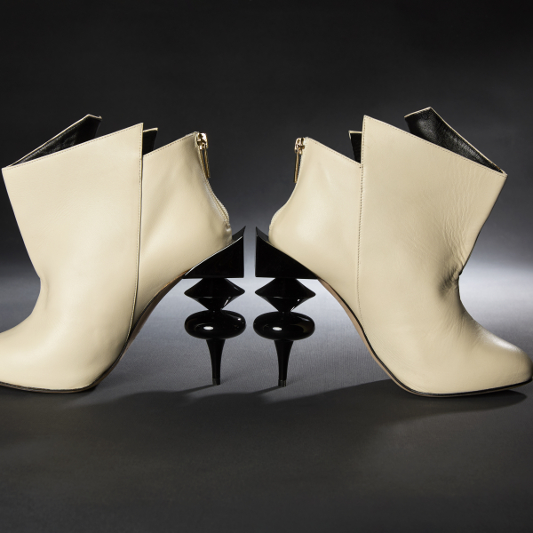  A dramatic photo of two high-heeled white leather boots with an intentionally jagged and angular top edge. The heels are formed of stacked geometric shapes and made of shiny black plastic, and the inside of the leather is black. The back of each shoe includes a visible silver zipper.