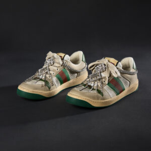 A pair of multicolored sneakers with chains of rhinestones draping over the center and the toe. The sneakers are tied with white laces and feature green and tan soles, with a mix of perforated white fabric, cream leather, and green and red striped ribbon details on the upper part of the shoe.