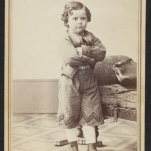 A sepia-toned portrait of a young male child posed in a photography studio with a patterned carpet. The boy has dark hair with ringlets, a pouty expression, and his arms crossed.