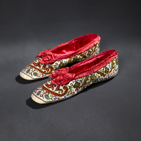 A pair of slippers with a white outer decorated with multicolored embroidery and a red inner with a ruffled edge and a bow over each front.