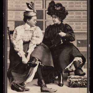 A tall rectangular card advertising Candee rubbers. Two women in long, ruffled clothing sit next to each other on a bench trying on shoes, with shelves of shoe boxes in the background.