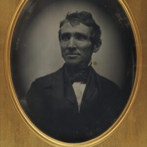 A black and white image of a white man from torso up dressed in a suit and bowtie, with mildly messy short hair. The image is oval and surrounded by a golden-colored frame.