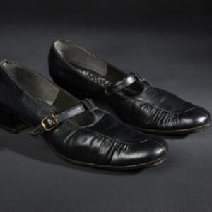 Two black shoes with crinkled leather-like fabric. The shoes each have a small block heel and a thin central buckled strap attached to a covered toe area.