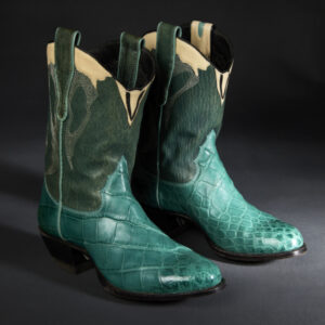 Two green and turquoise cowboy boots, with a snakeskin pattern on the turquoise lower portion and a hair texture and elaborate silver embroidery on the green upper portion. Near the tops of the boots, tan accents and two black and green finger loops.