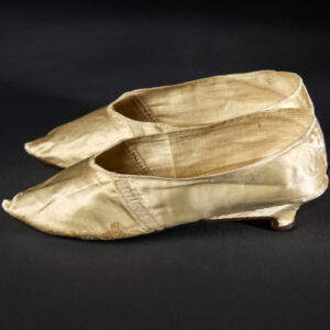 Two pointed flat shoes with a small heel shown in profile. The shoes are made of a tan-colored silky fabric, which shows signs of wear but also careful mending.