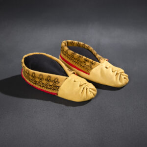 A pair of moccasins with a base of lighter-colored leather with a decorative wrinkled seam over the toe. The leather is folded over and decorated with black patterns and red edging around the ankle hole.