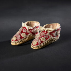 Small booties of red silk with elaborate clear beadwork and white silk ribbons in bows. They have a tan-colored sole with loops of beadwork where the sole meets the upper part of the shoe.