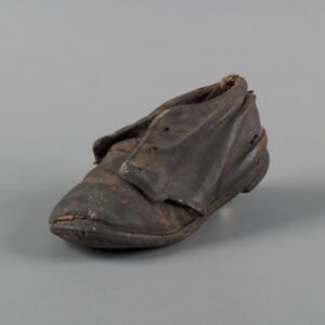A small, worn shoe of blackened leather. The shoe has holes for laces but no laces, and is slightly squished at the heel.