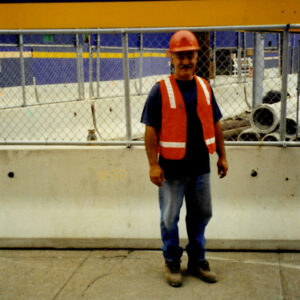 A man in a construction helmet, orange high-viz vest, a T-shirt, jeans, and worn construction boots stands in front of a construction site fence and concrete barrier. Buildings and a sign for I-93 are visible in the background.
