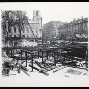 A view of a large construction site with trolleys on a road to the right and a church in the background. The construction site has been excavated many feet below street level and contains steel girders outlining what will become underground structures.