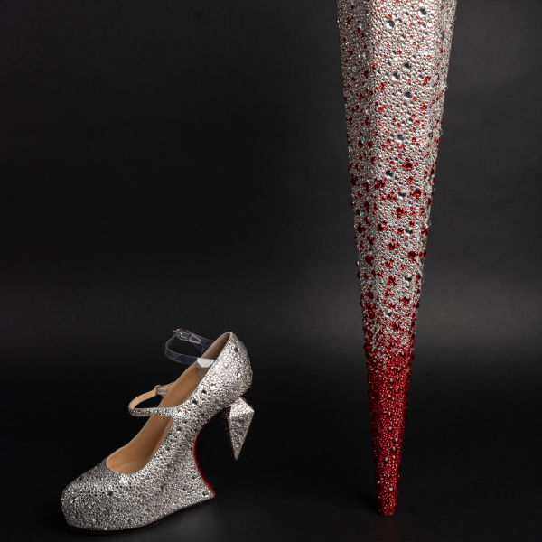 At left, a silver-rhinestoned cantilever shoe with a spike heel that doesn't touch the ground. The shoe has two straps, a visible rhinestoned strap and a transparent strap. At right, a spike prosthetic that transitions from silver rhinestones at the wider top to red rhinestones at the pointed bottom.