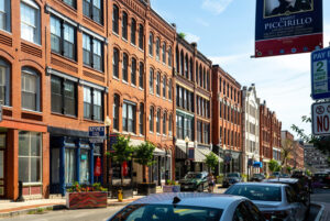 A street view looking at a long row of four-story brick buildings with decorate brickwork. A line of cars are parked along the street, and several shops, restaurants, and trees are visible along the sidewalk.