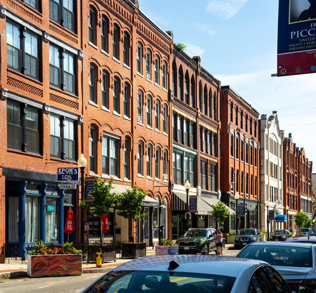 A street view looking at a long row of four-story brick buildings with decorate brickwork. A line of cars are parked along the street, and several shops, restaurants, and trees are visible along the sidewalk.