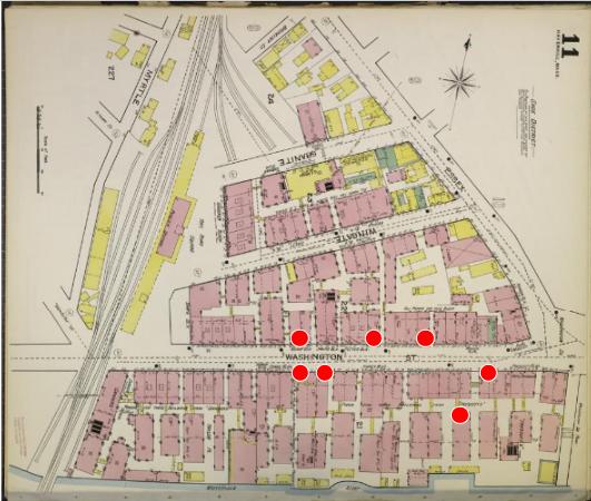 A map of showing a triangular area between the Merrimack River at the bottom, Essex Street at the right, and railroad tracks at the left. Washington Street runs parallel to the Merrimack River at the bottom of the map. Many pink (brick) buildings flank either side of Washington Street and fill the triangular area, with a few yellow (wood) buildings also visible. At the top right, text reads, “Shoe District.”