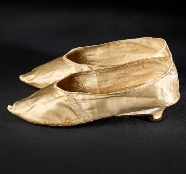 Two pointed flat shoes with a small heel shown in profile. The shoes are made of a tan-colored silky fabric, which shows signs of wear but also careful mending.