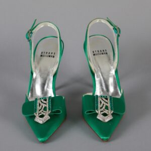 A pair of green and silver heels pointed toward the camera. There are green rhinestones on the silver brooch over the toe.
