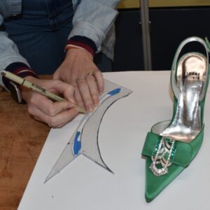 A pair of hands trace a shape in black marker onto a transparent piece of plastic next to a single green heel on a white workbench.