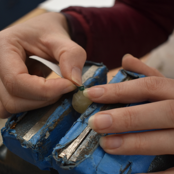 A very small green rhinestone mounted on a beeswax stick which is clamped upright in a vice. A pair of hands hold sandpaper to the surface of the rhinestone.