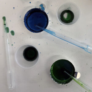 Overhead view of two plastic medicine cups and two dish-shaped aluminum trays filled with colored blue and green epoxy and several clear plastic pipettes.