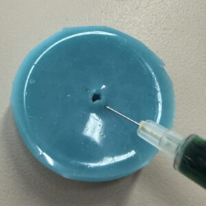 A close-up of a bright blue silicone mold with a syringe filled with green epoxy inserted into the silicone.