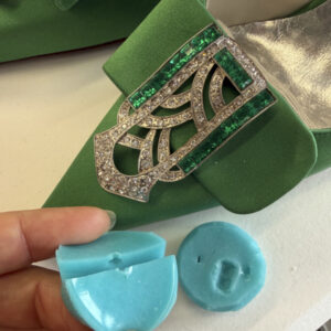 A close-up of a hand separating the two sides of a bright blue silicone mold to show the small gem-shaped cavity within. Another, slightly different mold sits to the right, and the green shoe is visible behind the molds.