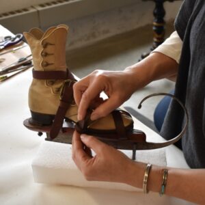 A conservator’s hands buckle new leather straps to hold a tan boot in place inside the antique ice skates.