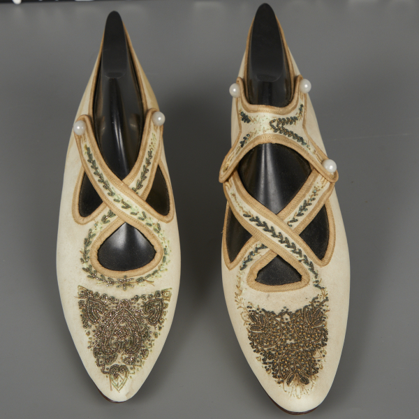 A pair of white leather shoes with black shoe forms inserted to hold their shape. The shoes are decorated with a range of decorative beadwork which has begun to corrode and is missing in some areas.
