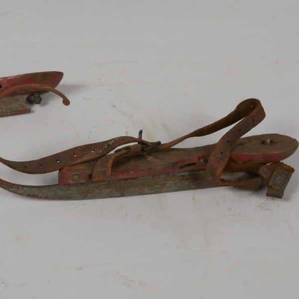 A pair of antique ice skates made of a long blade, a wooden platform, and leather straps with buckles.