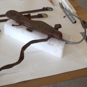 One antique ice skate with its blade wedged into white foam to stand it upright. Its leather straps are unwound and laid out next to the skate. One piece is broken off.