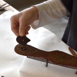 A conservator’s hand holds a piece of flat shim with a metal stud over the sharp-looking spike extending from the top of the wooden skate.