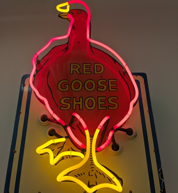 A lit neon sign in the shape of a red goose outlined in red and yellow neon. Text on the backing board reads, “Red Goose Shoes.”