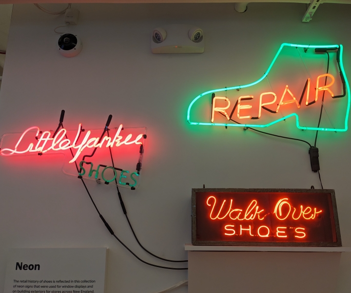Three lit neon signs arranged on a white wall. The left sign is pink and green and reads, Little Yankee Shoes. The top right sign reads REPAIR in capital red letters, outlined in the shape of a green boot. The bottom right sign reads Walk Over Shoes in bright red neon.
