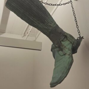 A large green-colored copper sign hangs from a wall. The sign is a hollow three-dimensional shoe with a bow at the ankle and a long stocking that extends upward.