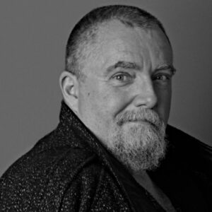 A white man with short-shorn hair and a full moustache and beard looks confidently at the camera in a three-quarters black and white view.