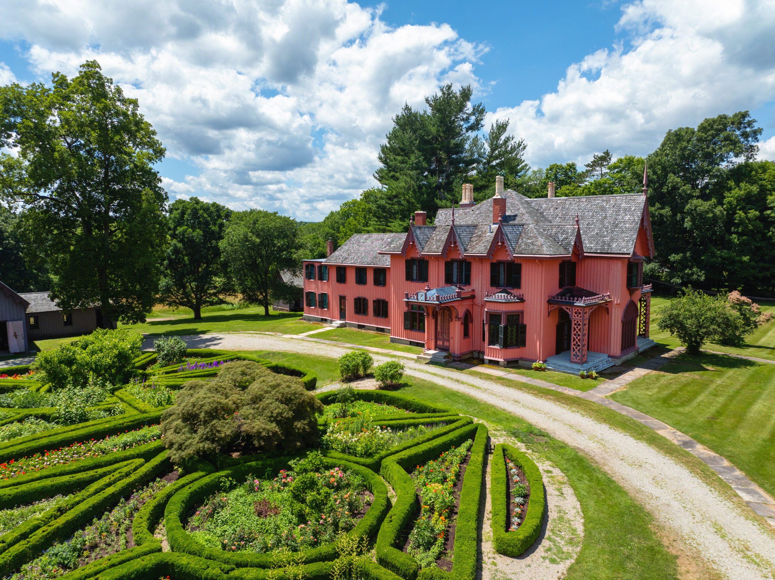 Exterior view of Roseland Cottage, showcasing a pink painted house in the background with a large garden of sculpted hedges and planting beds in the foreground.