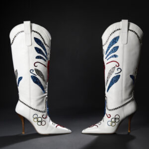 Two knee-high high-heeled white cowboy boots bedazzled in red, white, and blue rhinestone patterns. The Olympic rings are visible along the outer edge of each foot.