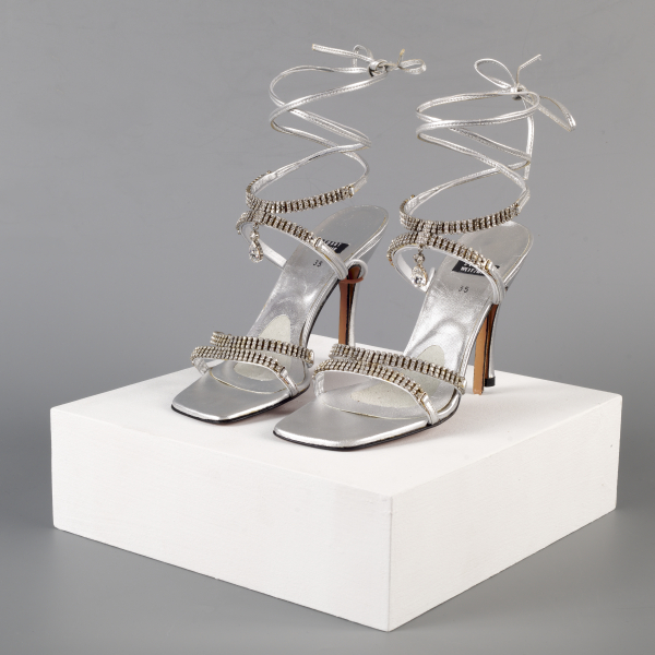 A pair of silver strappy rhinestoned heels. Each shoe has two conjoined rhinestone straps over the toe, and two conjoined rhinestone straps and a dangling pendant over the front ankle , with thin silver straps held up by wire winding up where there would be an ankle and lower leg of a wearer.