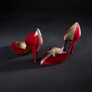 A pair of red satin pumps, each with a large clear rhinestone arrow. The left heel has an arrow on the toe pointing to the interior of the shoe, while the right heel has an arrow on the heel pointing up towards a wearer’s leg.