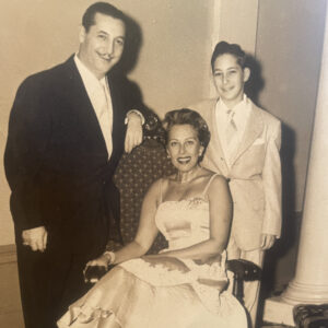A family photo with a mustache-wearing man in a suit at left, a woman in a white dress seated at center, and a boy wearing a light-colored suit at right. They are smiling for the photo.