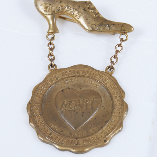 A brass brooch of a high-heeled shoe with a medal suspended from it by two chains. The medal reads, “Hitch your heart to Haverhill” at the center.