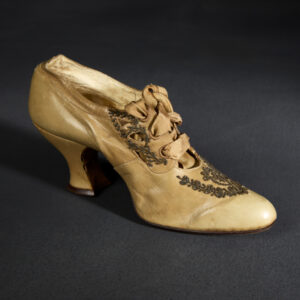 A single enclosed high-heeled shoe of light tan leather. The front features decorative beadwork and several cut-outs where a silk ribbon is laced through.