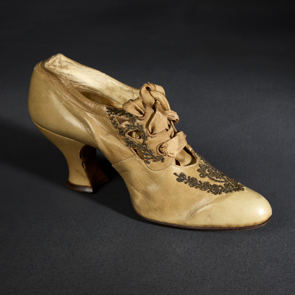 A single enclosed high-heeled shoe of light tan leather. The front features decorative beadwork and several cut-outs where a silk ribbon is laced through.