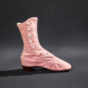 A high pink silk boot with a low black heel. Eight white glass buttons fasten a silk flap over the ankle of the shoe.